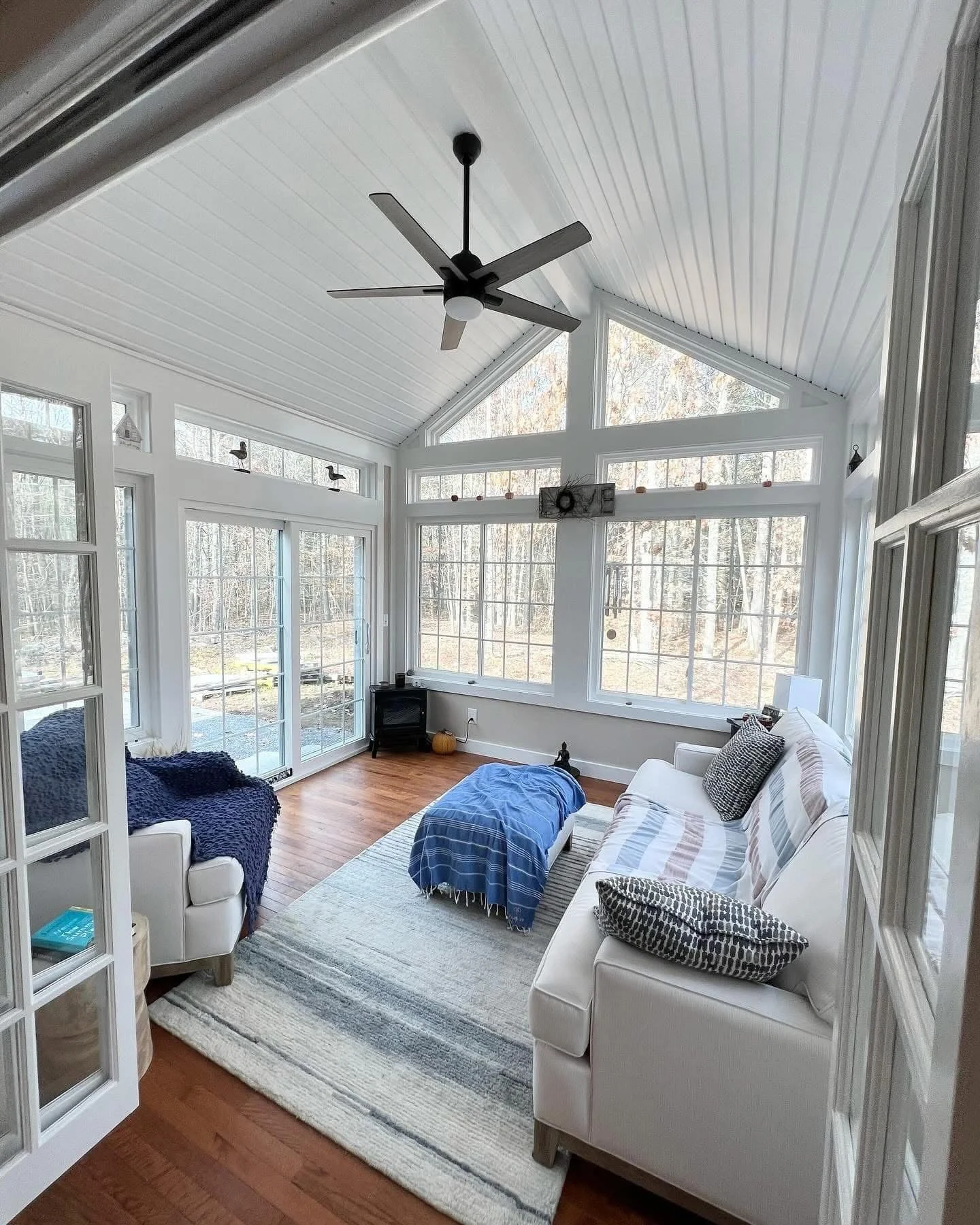 Sunroom with wood flooring, white furniture, large windows, vaulted ceiling, ceiling fan, and outdoor view.