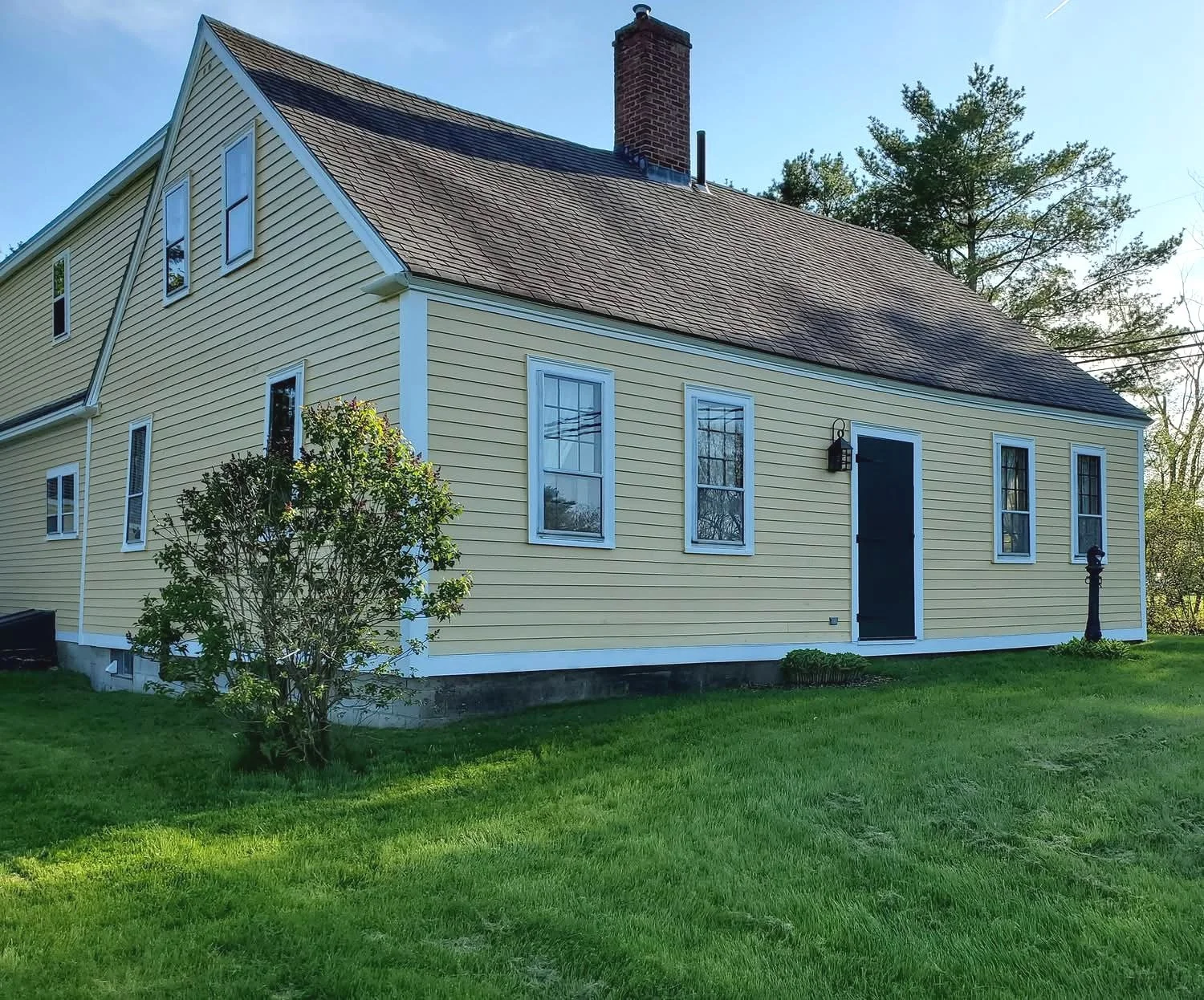 A yellow house with white trim and a brick chimney in a grassy yard, surrounded by trees, on a clear day.