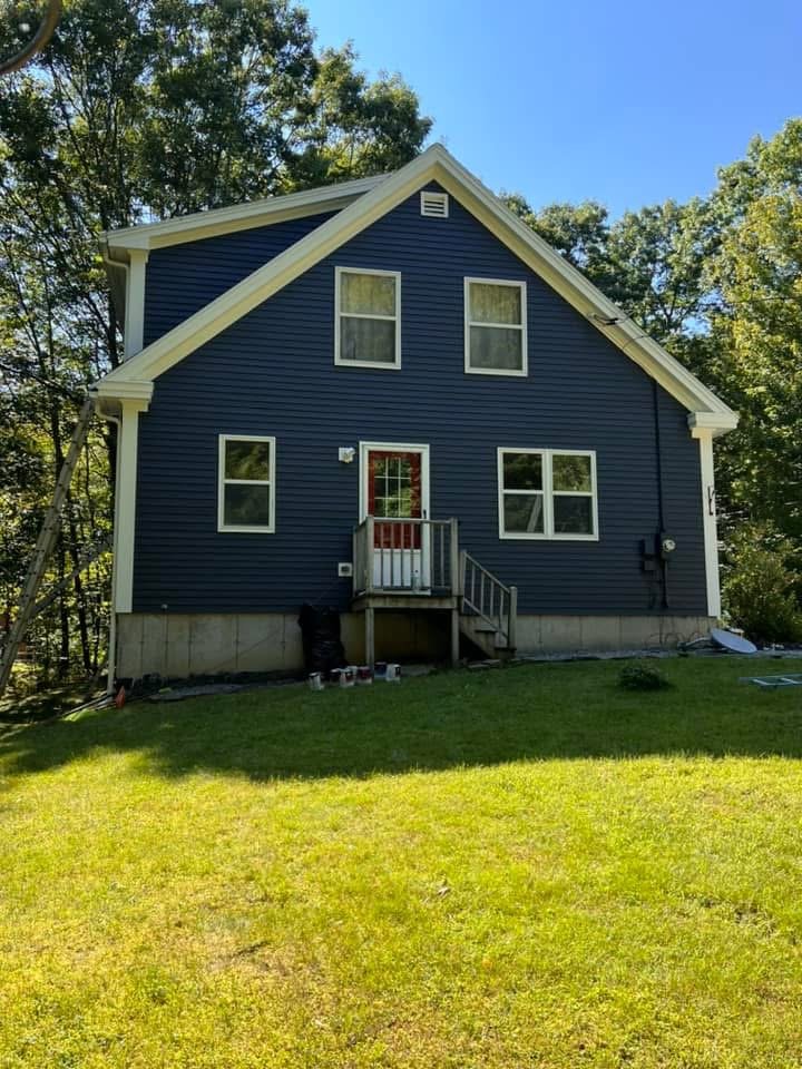 A two-story blue house with white trim, several windows, a small deck with stairs, and surrounded by trees and a grassy yard.
