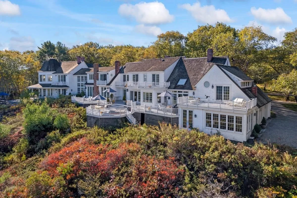 Large white residential house with multiple decks and large windows, surrounded by trees with autumn foliage, under a partly cloudy sky.