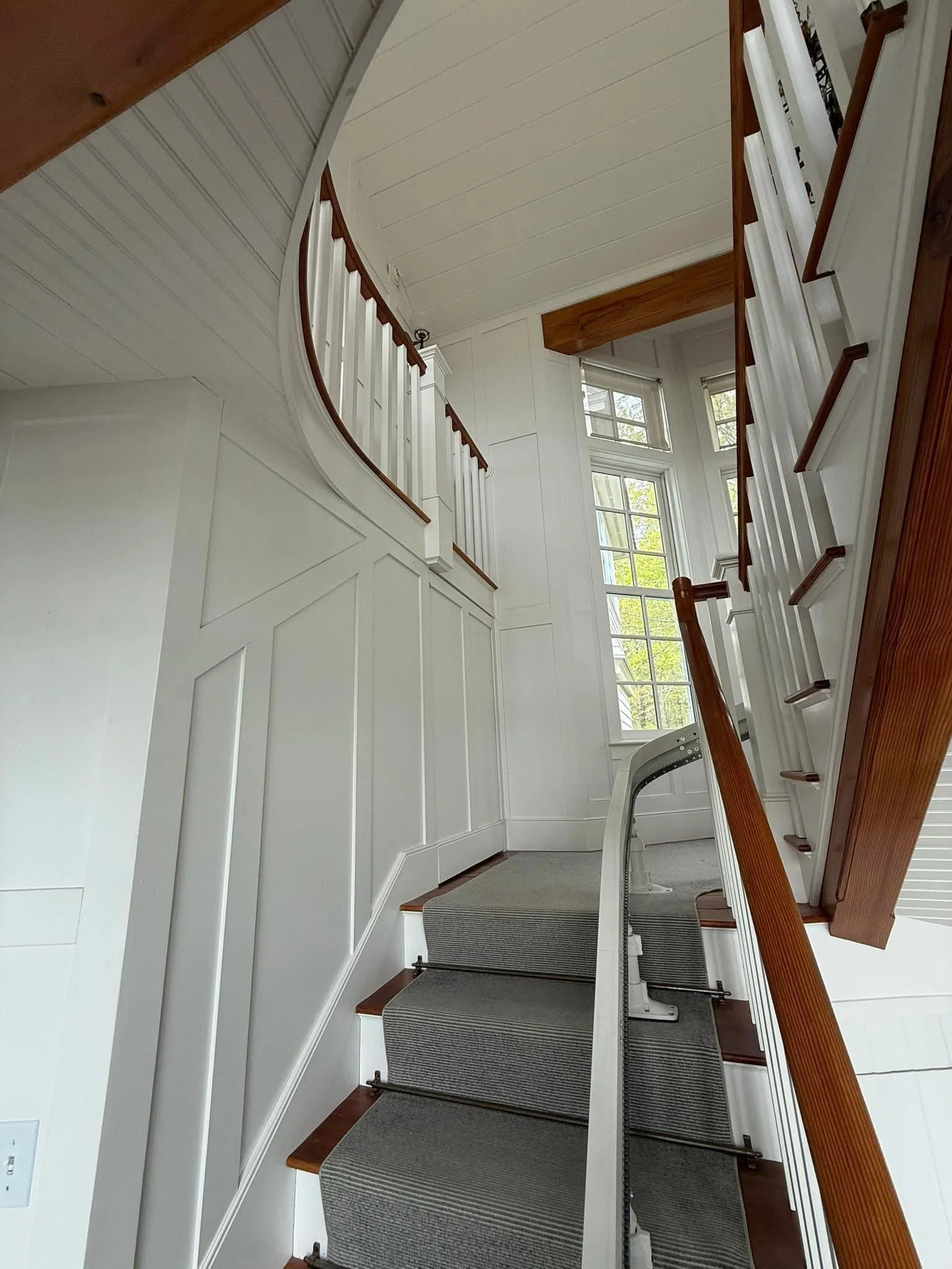 Interior view of a staircase with carpeted steps, white wooden panel walls, large windows, and a curved balcony railing. Spring or summer foliage visible outside the windows.