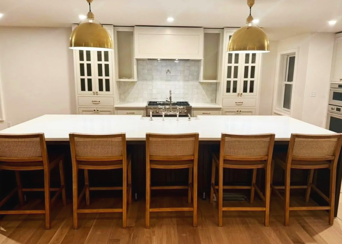 Modern kitchen with white cabinetry, marble backsplash, and a large white countertop island with six wooden bar stools. Two gold pendant lights hang above the island.