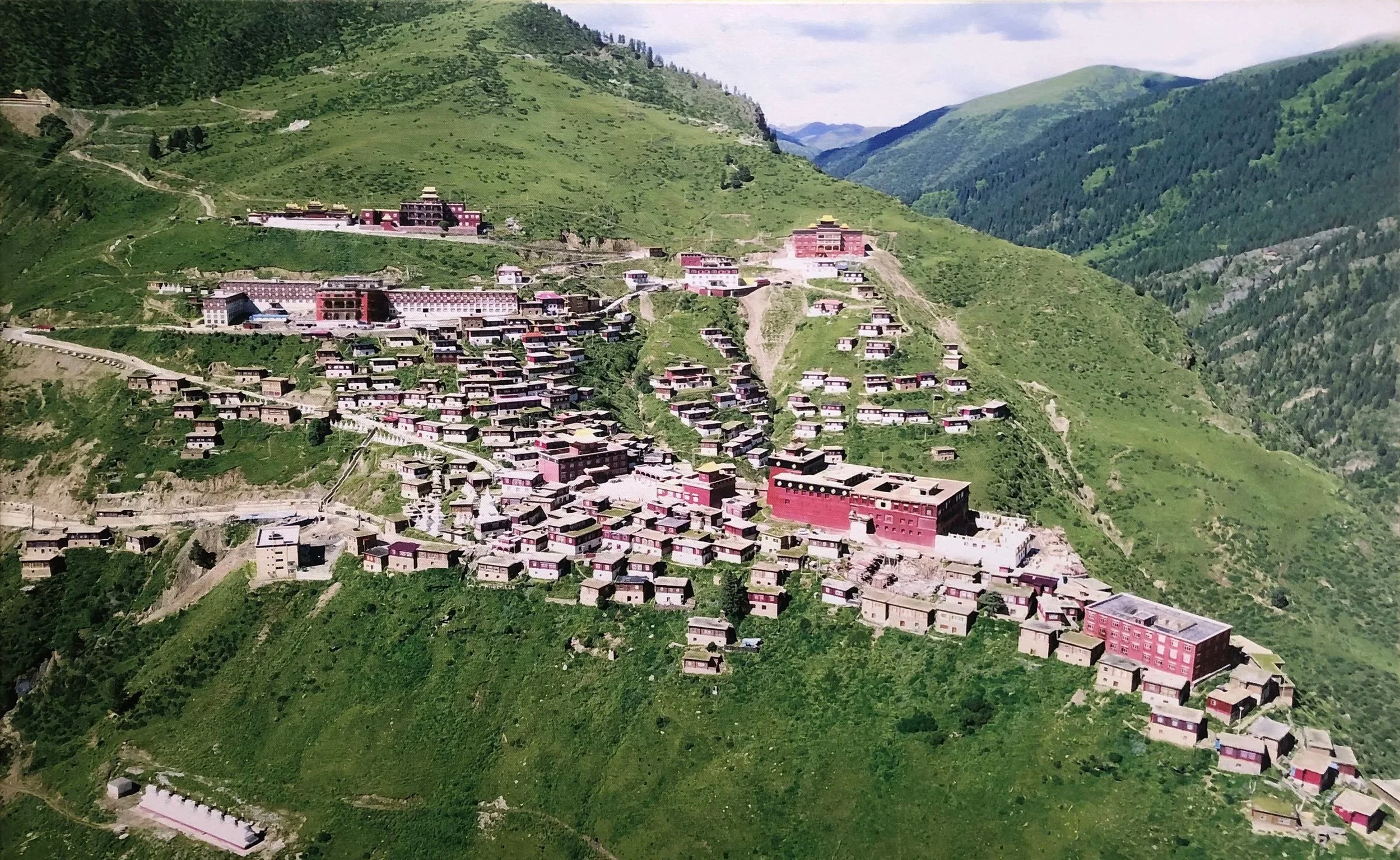 A mountainous hillside village with numerous small houses and larger buildings, surrounded by green grass and trees, with winding paths leading through the village.