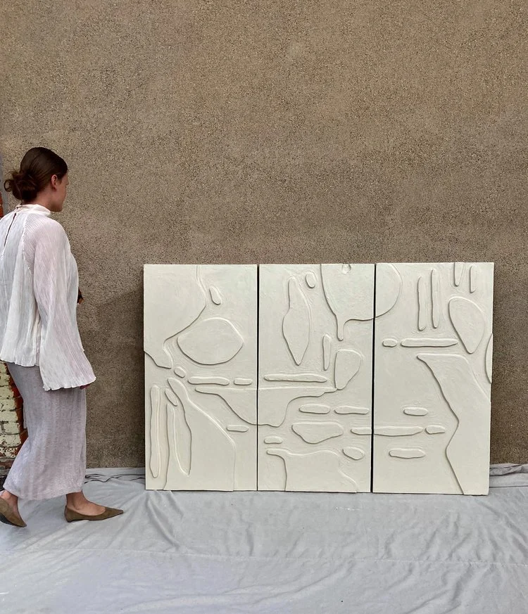 A woman in a white blouse and gray skirt standing next to three white abstract textured panels on a beige wall.