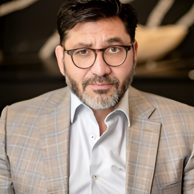 A man with dark hair, glasses, and a beard wearing a light plaid blazer and white shirt.
