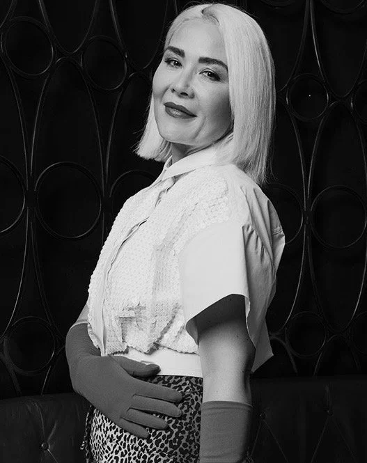 Black and white photo of a woman with shoulder-length blond hair, wearing gloves, a textured top, and animal print pants, smiling with a dark patterned background.