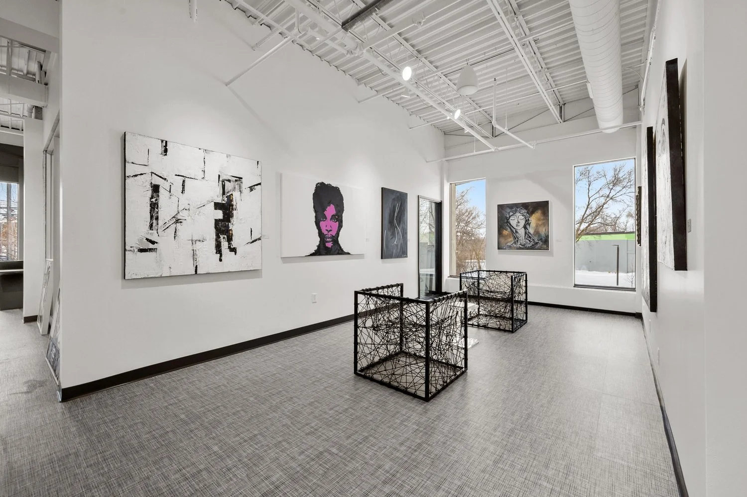 Contemporary art gallery with white walls, several paintings including abstract, portrait, and figurative works, black wire sculptures on the floor, and large windows showing a view of trees outside.