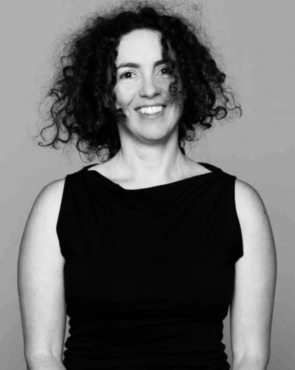 Black and white portrait of a woman with curly hair smiling, wearing a sleeveless black top against a plain background.