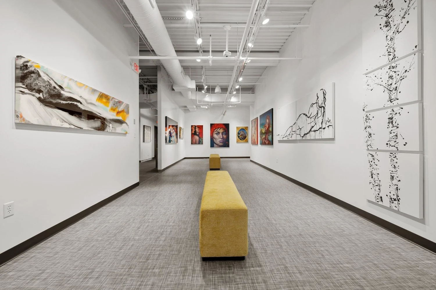 Art gallery with white walls displaying various paintings, yellow benches in the center, and a carpeted floor.