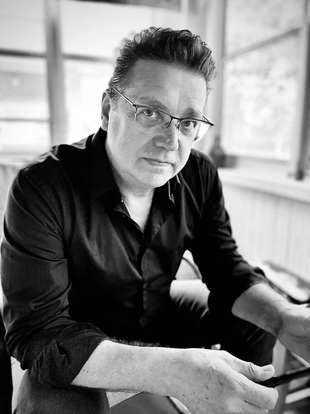 A black-and-white portrait of a middle-aged man with glasses, wearing a dark shirt, sitting indoors near a window with a blurred background.
