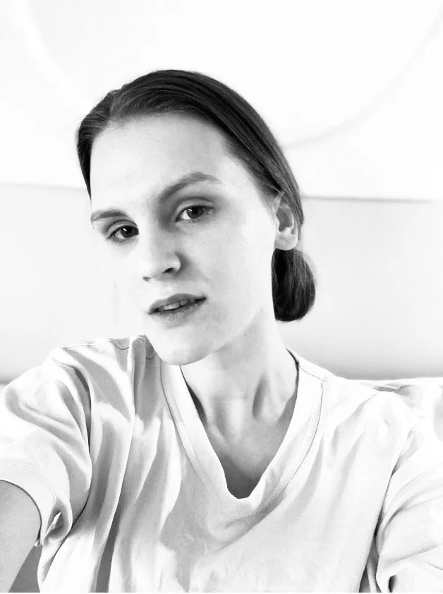 Black and white photo of a woman with dark hair pulled back, wearing a light-colored shirt, sitting against a neutral background.