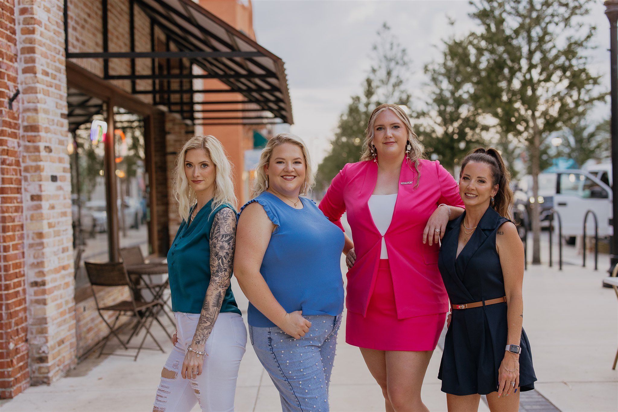 Four women standing on a sidewalk outside a brick building, smiling and posing for a photo.