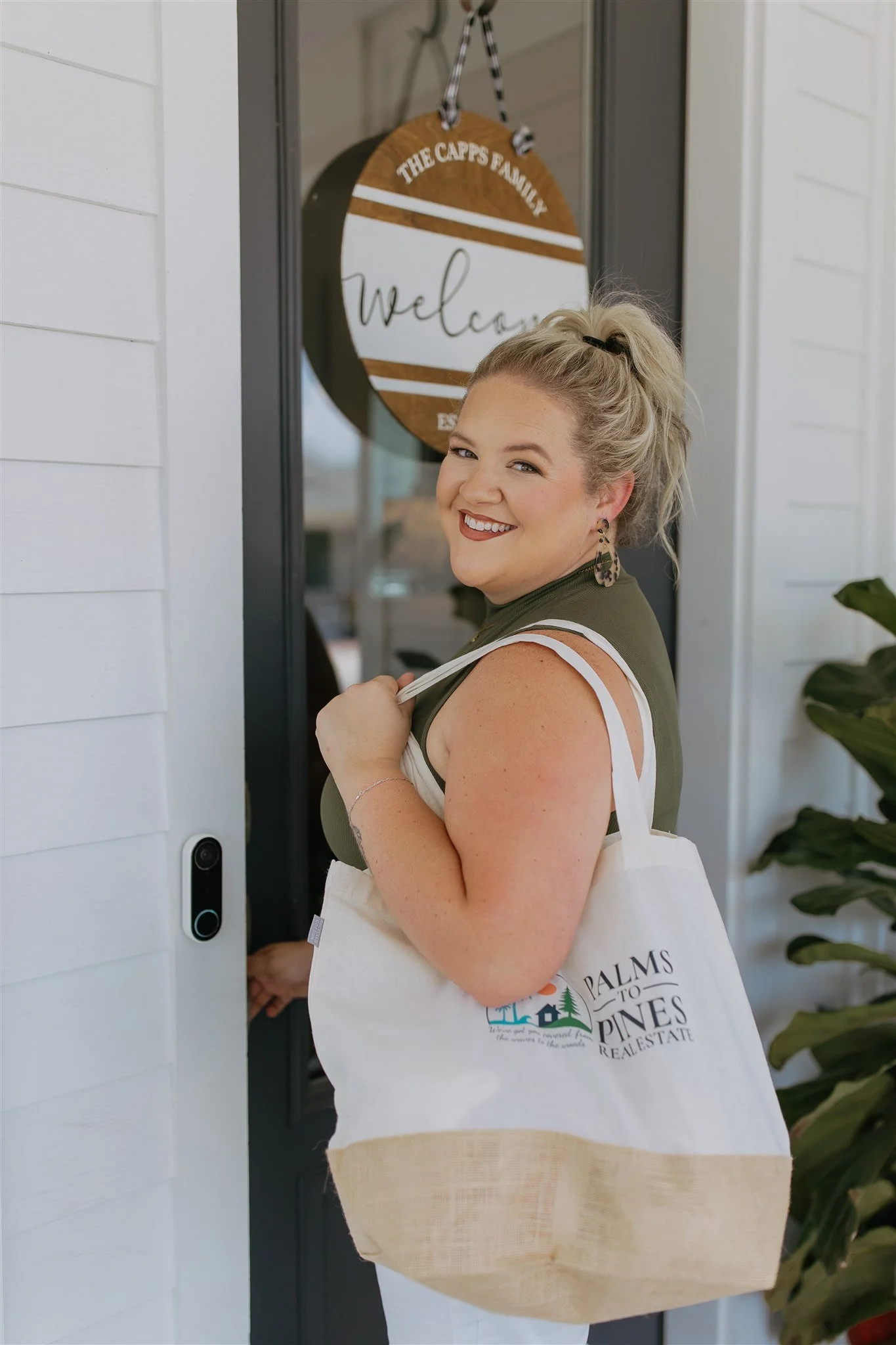 A smiling woman stands at her front door holding a tote bag. The door has a round wooden welcome sign that says "The Capps Family" and "Welcome." She has blonde hair in a ponytail, is wearing earrings, a sleeveless green top, and has a tattoo on her arm.