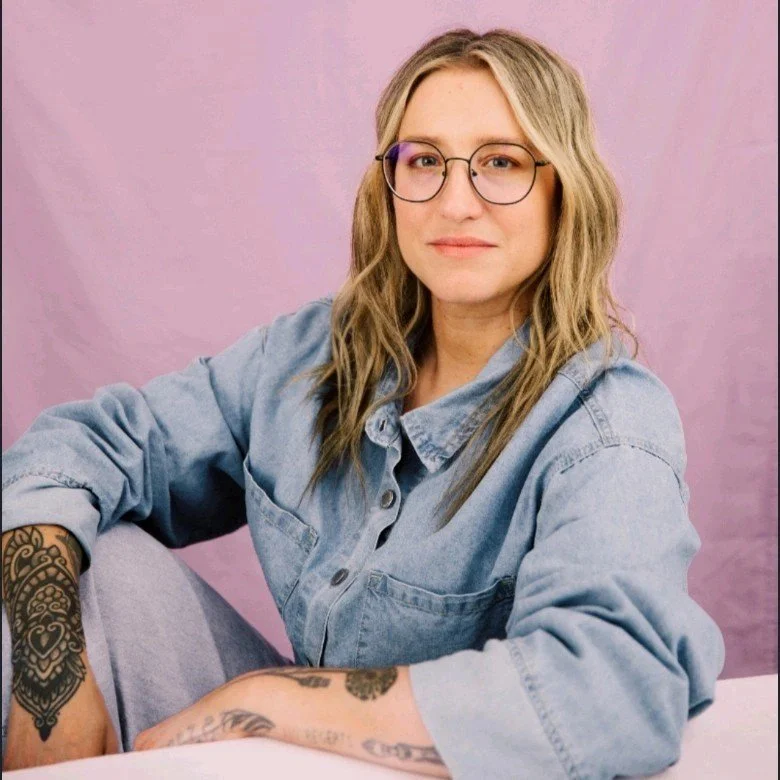 A woman with wavy blonde hair, glasses, and tattoos on her arm, sitting against a pink background, wearing a light blue denim jacket.