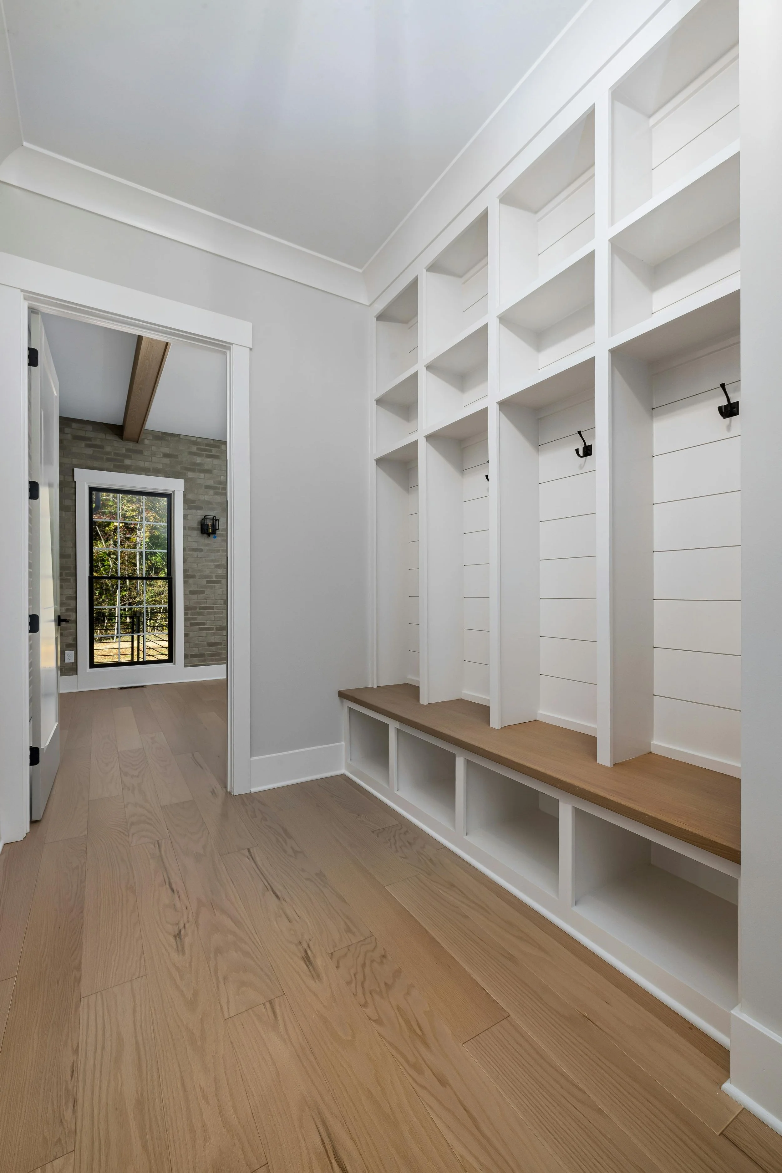 Empty hallway with built-in white storage unit with hooks and a wooden bench, light wooden flooring, and a doorway leading to a room with a brick wall and windows showing trees outside.
