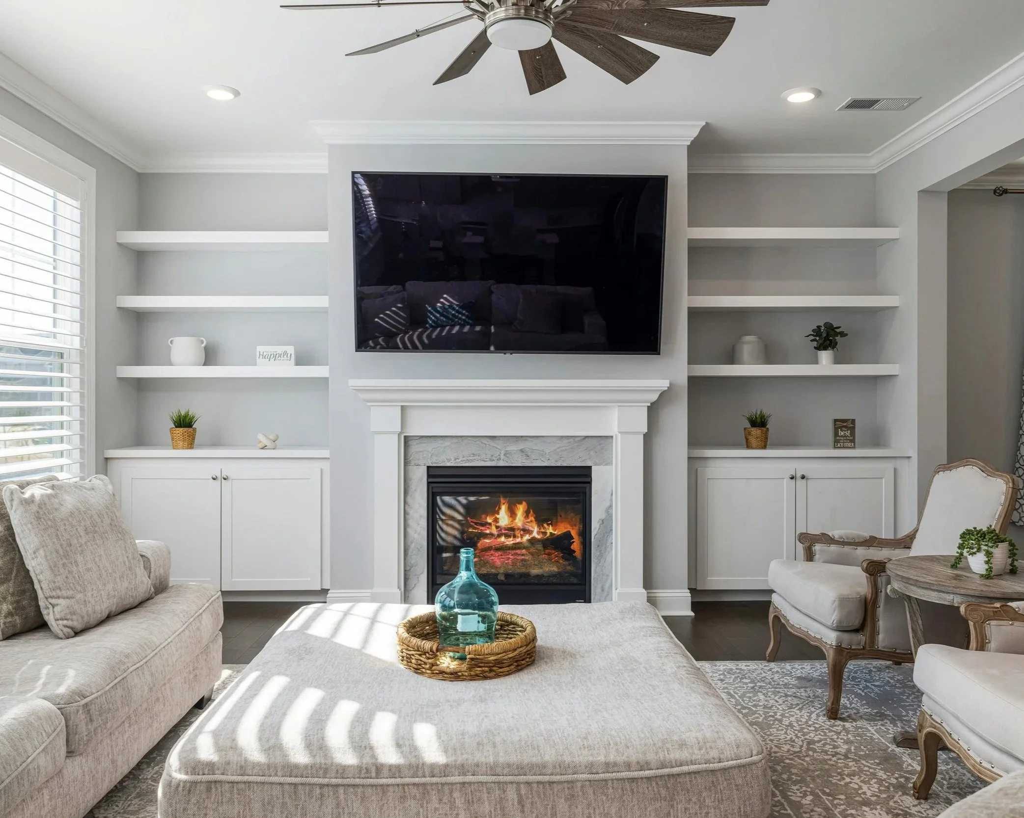 Living room with a fireplace, a wall-mounted TV above it, beige sofas, a white armchair, built-in white shelves with decorative items, and a ceiling fan.