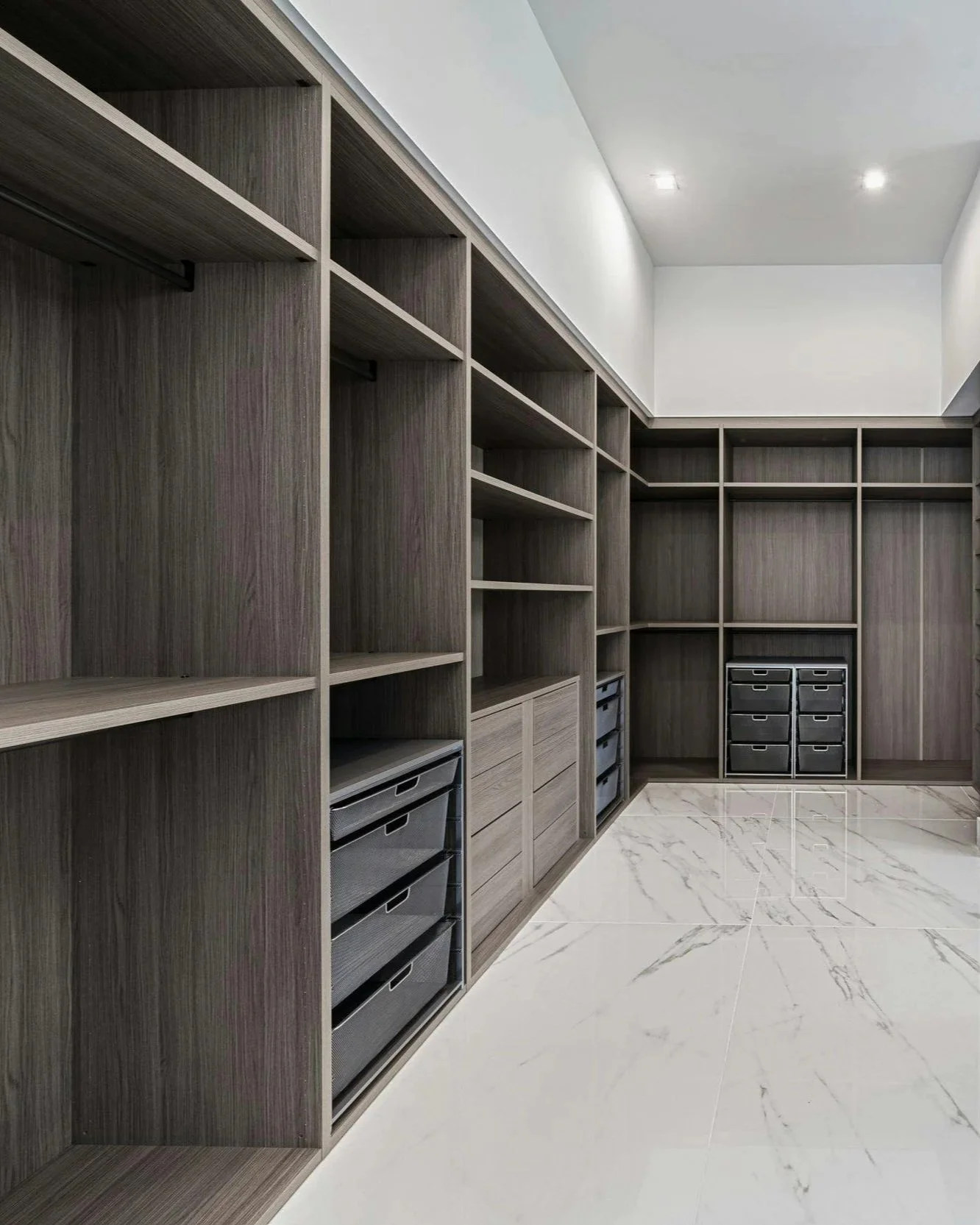 Empty walk-in closet with wooden shelves, drawers, and black storage bins, with white marble tile floor and recessed ceiling lights.