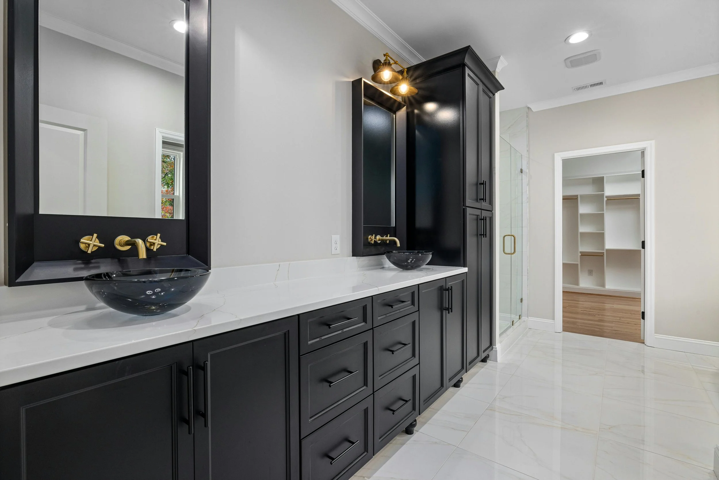 Bathroom with black cabinets, white countertop, two black vessel sinks with gold fixtures, large mirror, and glass shower door.