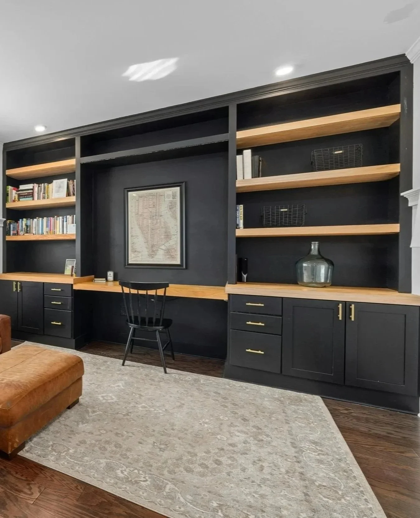 A black built-in bookshelf with wooden shelves, containing books, decorative items, and a framed map, with a desk and chair in front.