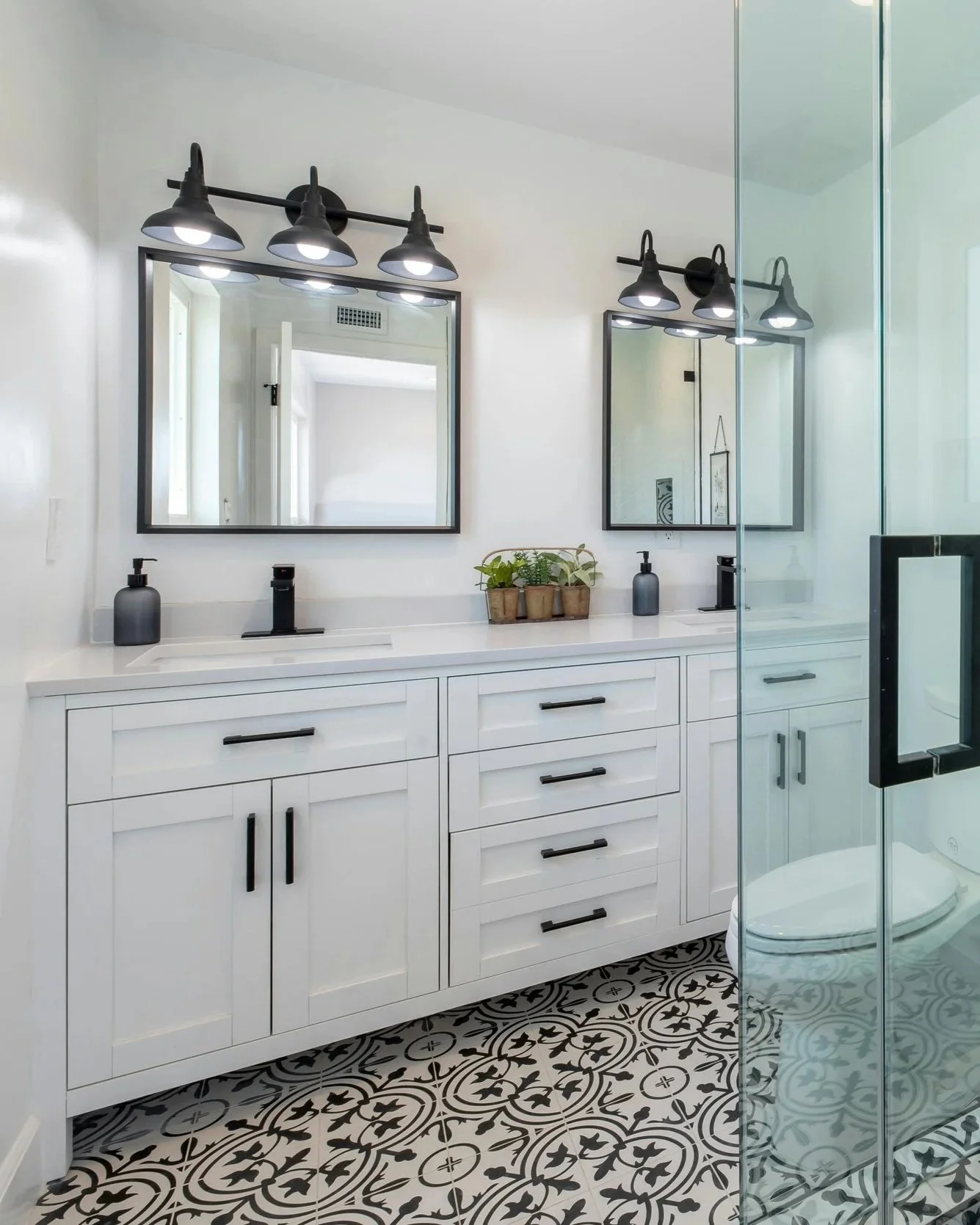 Modern bathroom with double vanity, black fixtures, black frameless mirrors, stylish black light fixtures, potted plants, patterned black and white tile floor, and glass shower enclosure.
