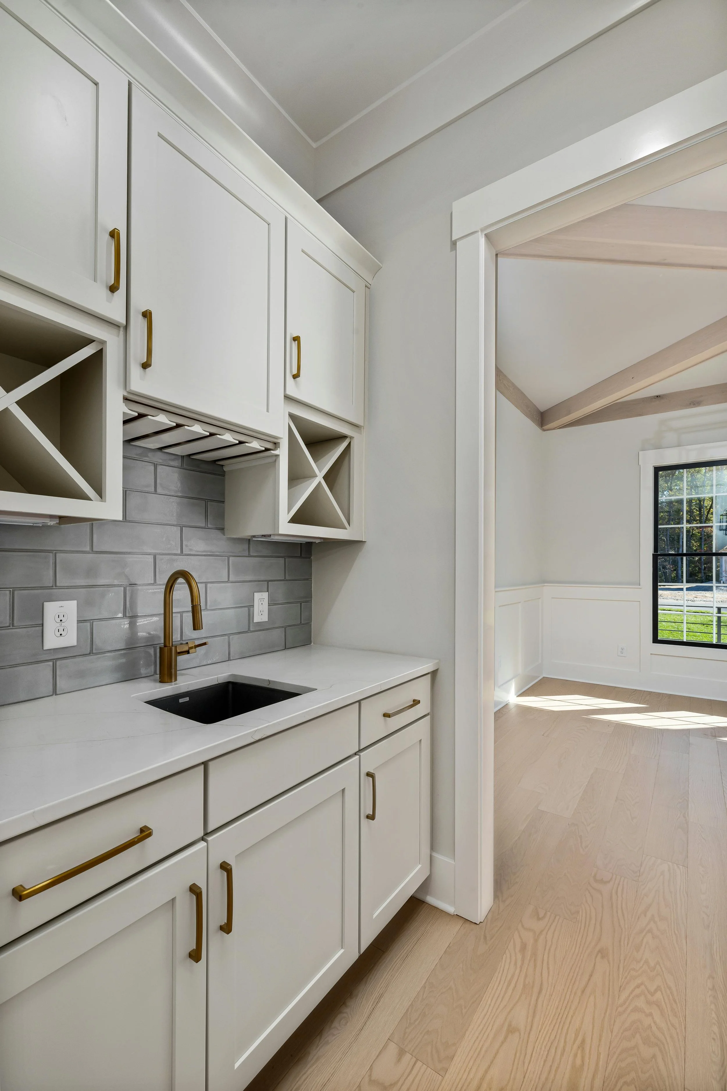 Interior view of a kitchenette with white cabinets, gold handles, a black sink, gold faucet, gray subway tile backsplash, and open wine racks, adjacent to a bright room with large windows and light wood flooring.