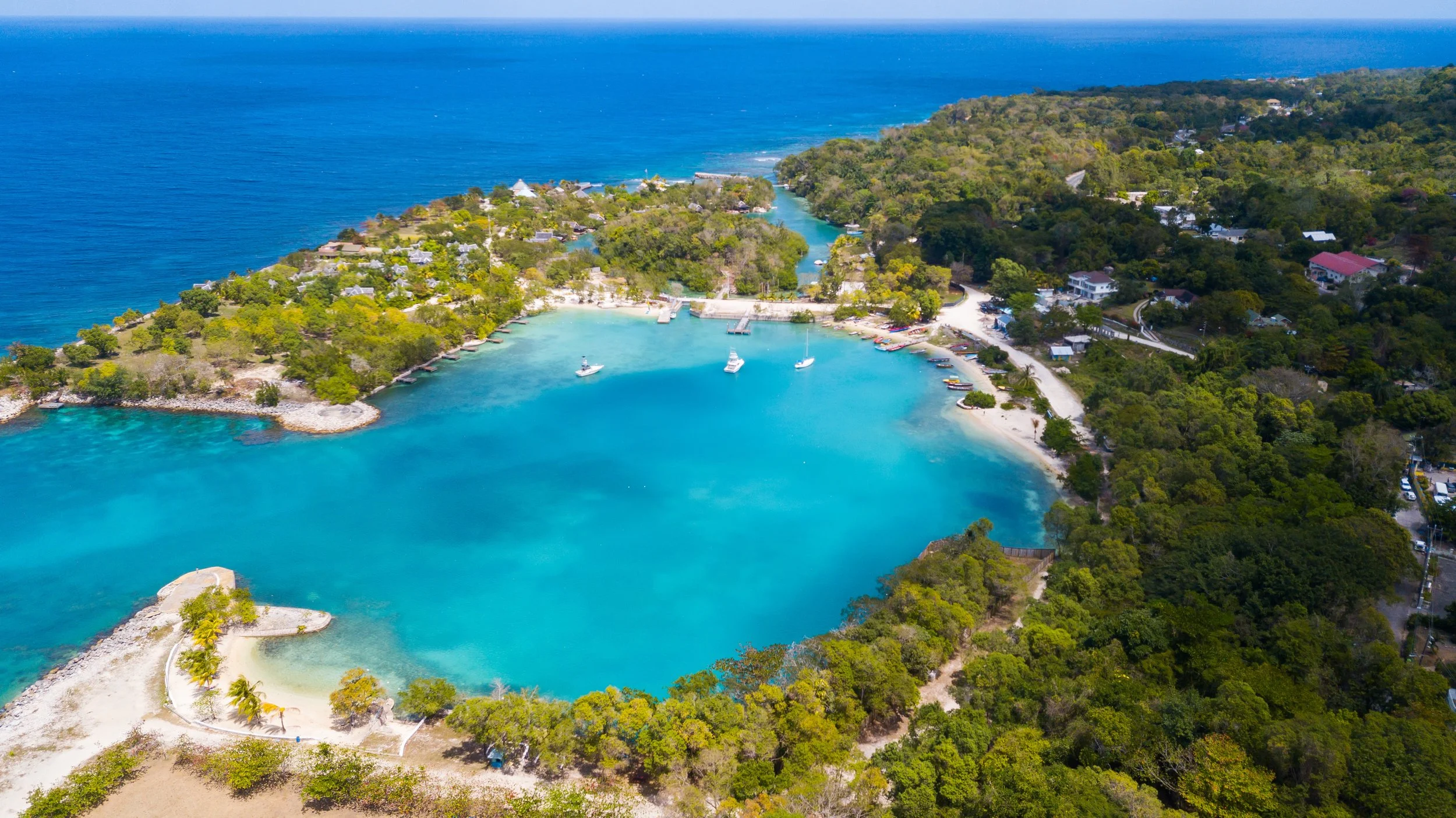 Aerial view of a tropical bay with turquoise waters, boats anchored near the shore, sandy beaches, lush green trees, and hillside houses. The ocean extends into the horizon.