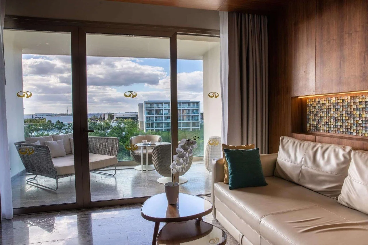 Living room with a beige leather sofa, dark pillows, and a small round coffee table holding a white orchid. Large glass sliding door leads to a balcony with outdoor seating, overlooking a cityscape with trees and modern buildings under partly cloudy skies.
