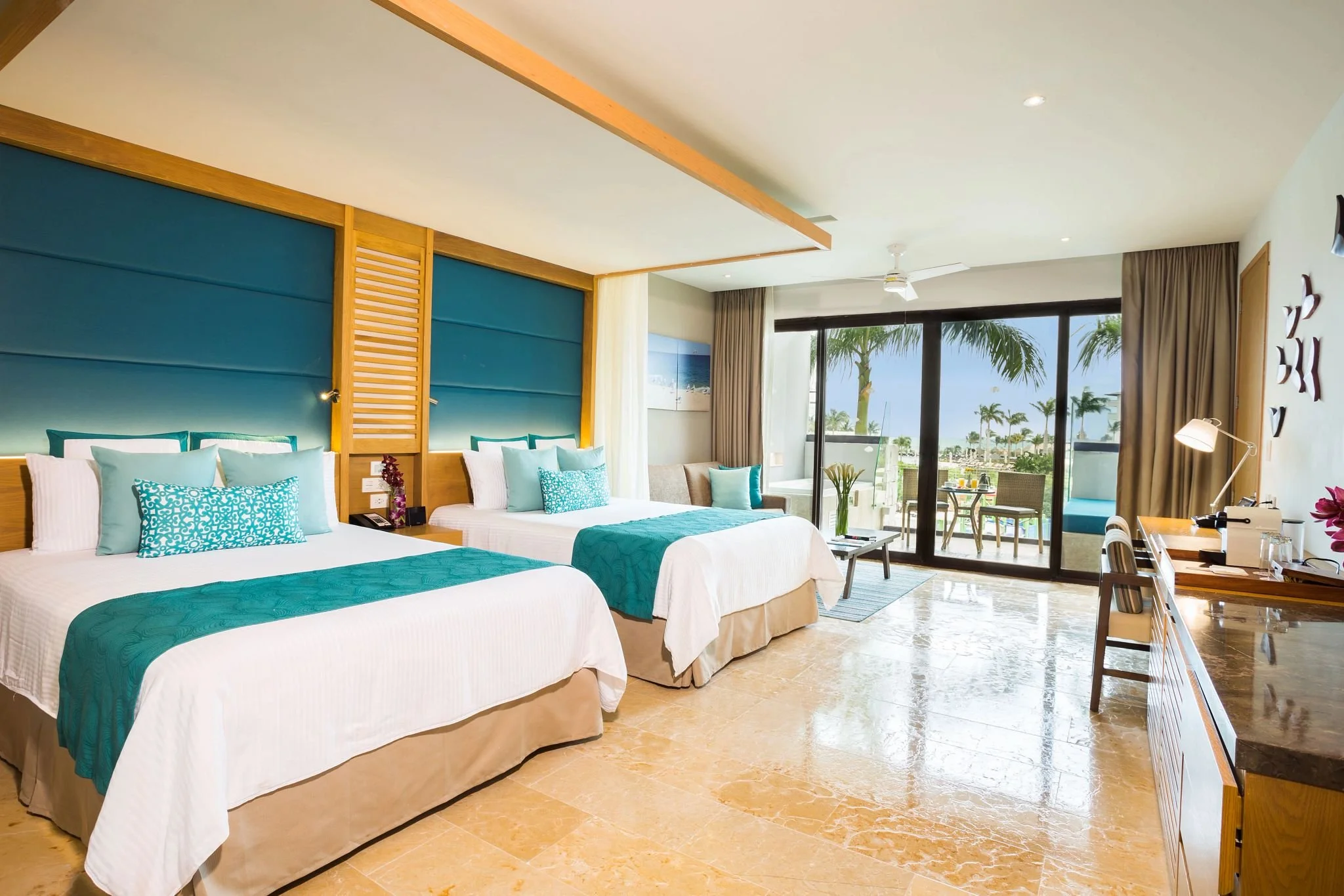 Hotel room with two beds, teal accents, a balcony with outdoor seating, and palm trees visible outside.