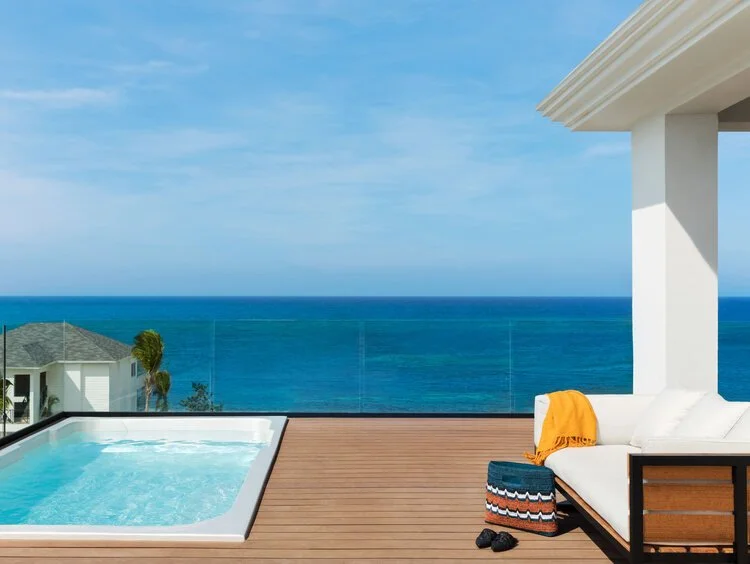 A balcony overlooking the ocean with a hot tub, white outdoor furniture with pillows, an orange towel, and a small round table with a striped cover.