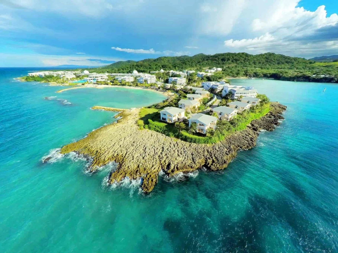Aerial view of a tropical island with rocky coastline, white buildings, and green hills in the background, surrounded by clear turquoise ocean water.