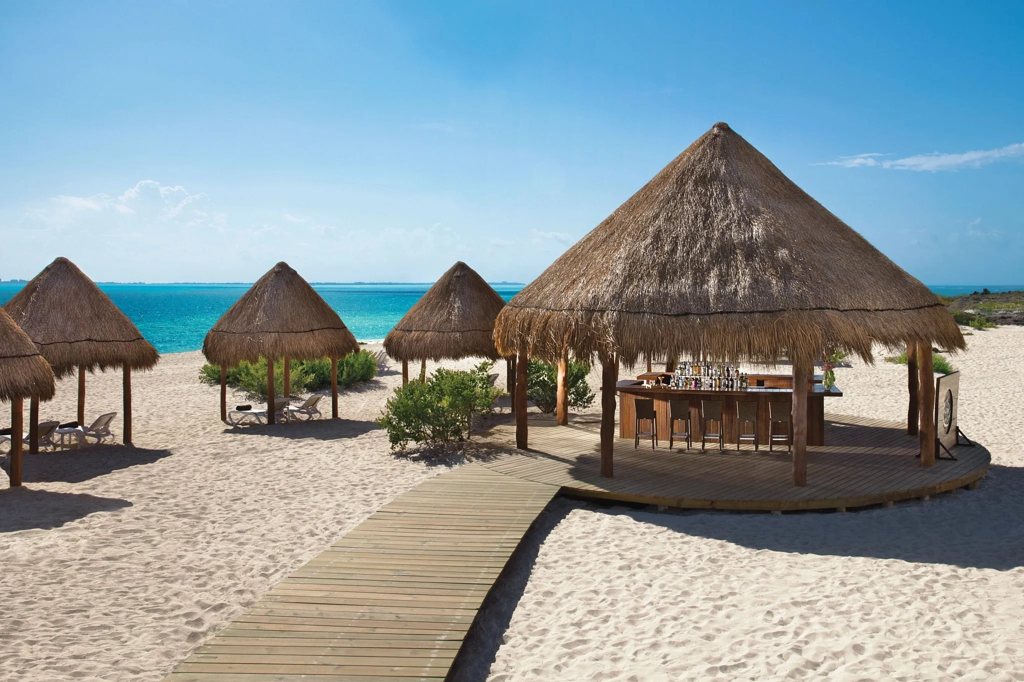 Beach scene with six thatched-roof palapas, a wooden walkway, lounge chairs, green bushes, and the ocean in the background under a blue sky.