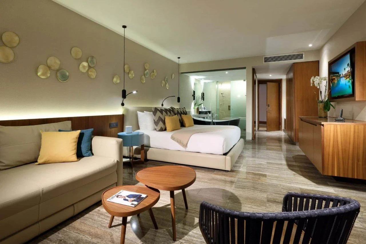 Modern hotel room with a bed, sofa, coffee tables, TV, and a view of the bathroom with a bathtub and shower.