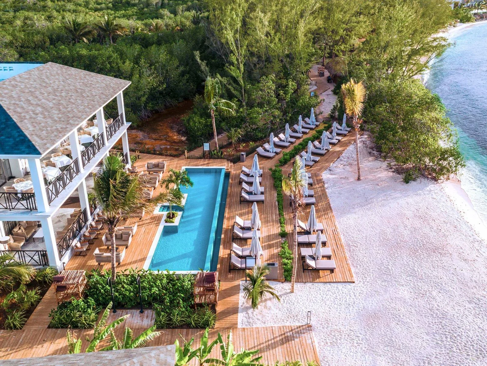 Aerial view of a beachfront resort with a swimming pool, sun loungers with umbrellas, and a sandy beach bordered by greenery and trees.