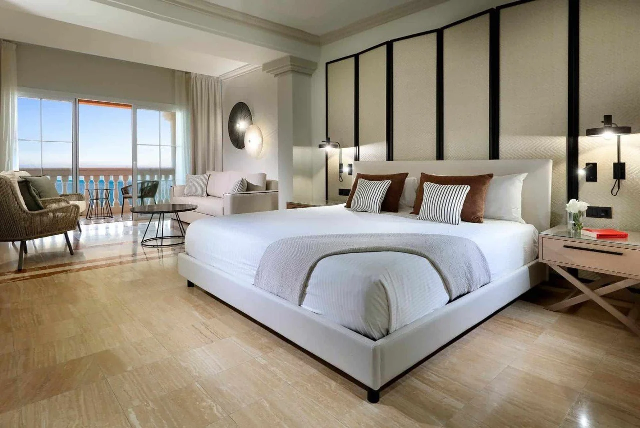 A modern hotel room featuring a large bed with multiple pillows, a beige and brown color theme, a bedside table with flowers and a red book, a sitting area with two chairs and a coffee table, and sliding glass doors leading to a balcony with ocean view.