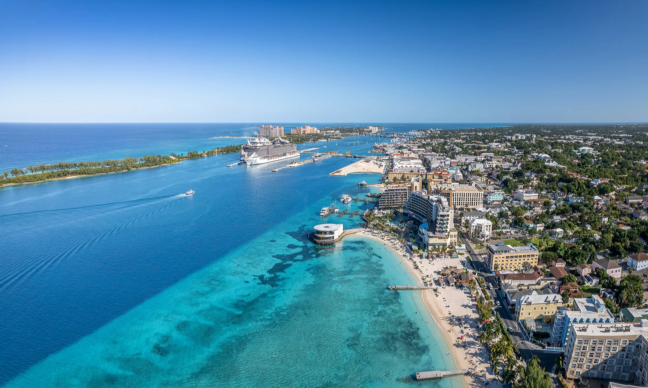 Aerial view of a coastal city with a harbor, cruise ships, beaches, and high-rise buildings along the shoreline, with clear blue water and a bright sky.