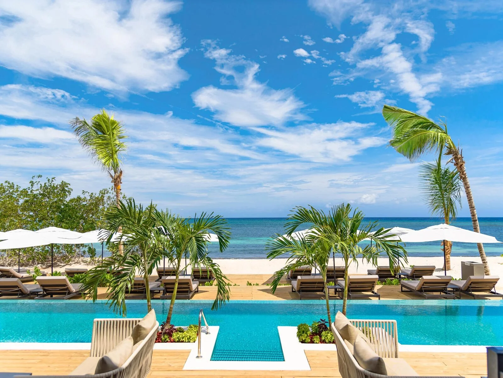 A tropical beach resort with a swimming pool, lounge chairs with umbrellas, palm trees, and the ocean in the background under a partly cloudy sky.