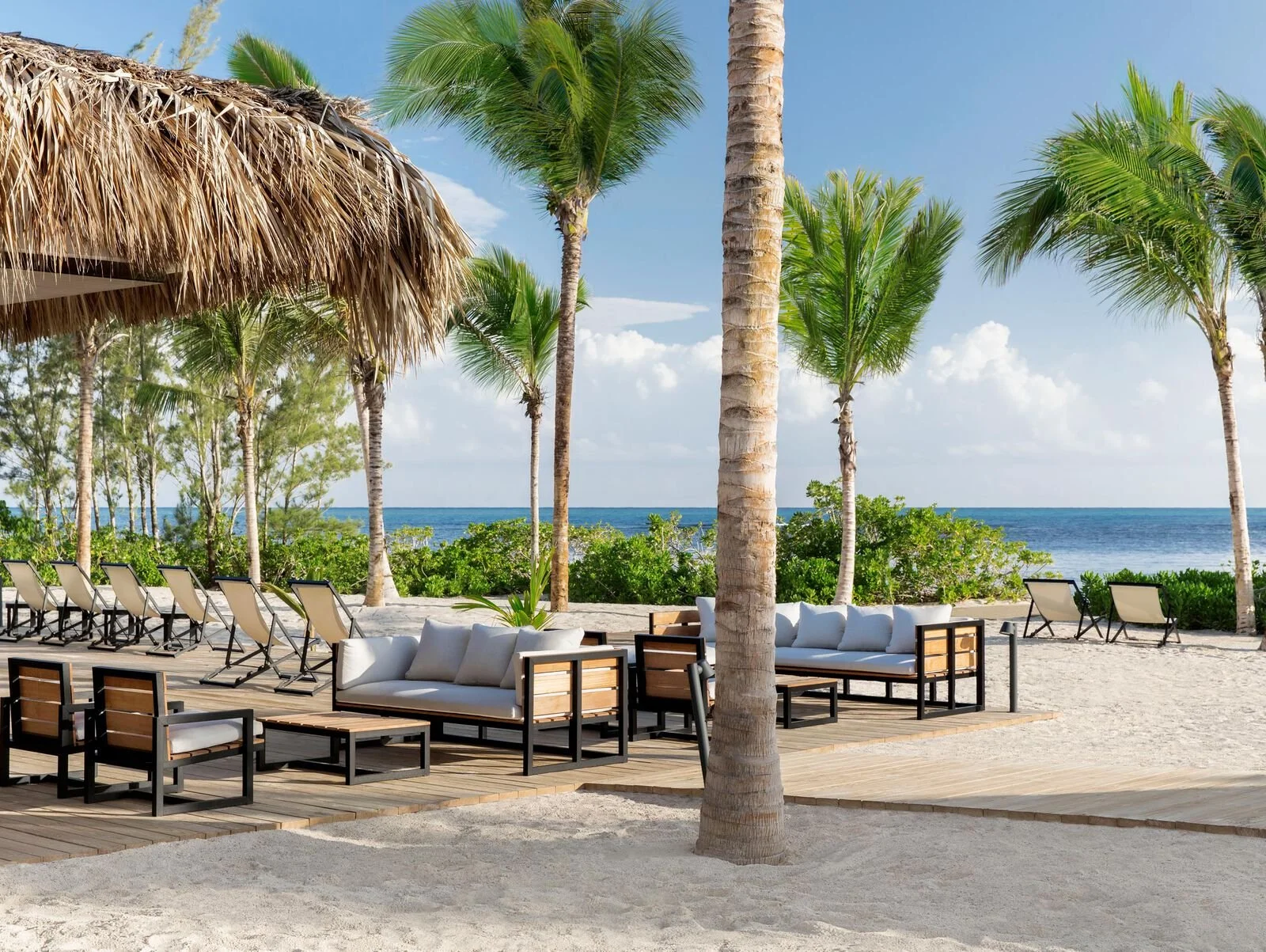 Beachside lounge area with white sofas and chairs on wooden deck, surrounded by palm trees, overlooking the ocean under a partly cloudy blue sky.