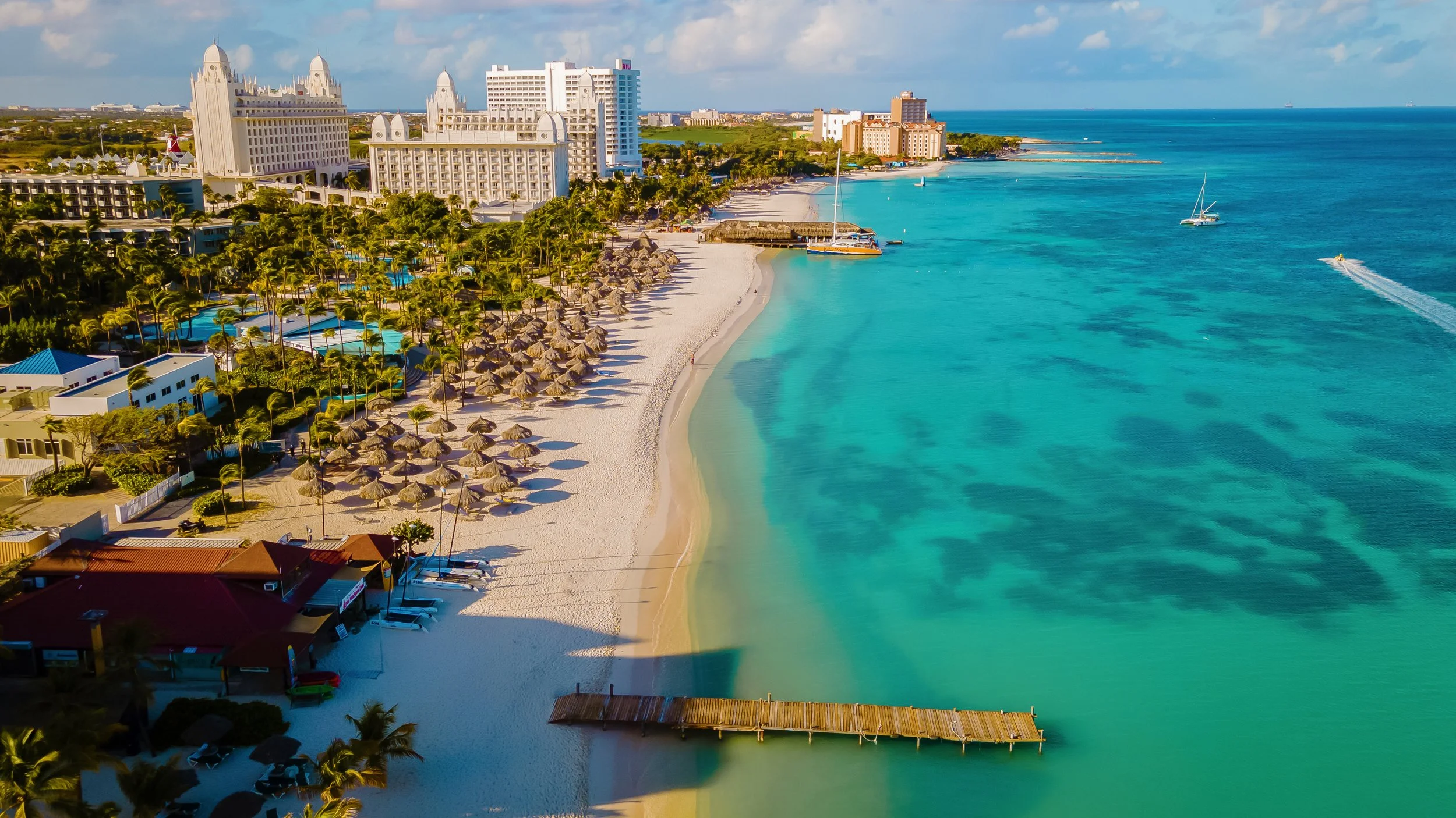 Aerial view of a tropical beach with white sand, turquoise water, and thatched umbrellas. High-rise hotels and resort buildings line the coast, with boats and sailboats on the water.