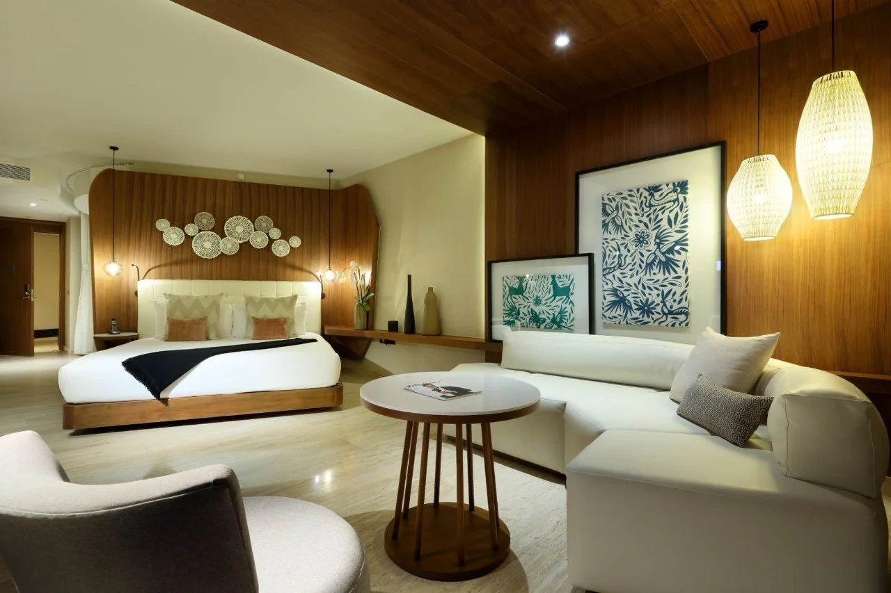 Modern hotel room with a king-sized bed, white sofa, armchair, wooden wall decor, and hanging pendant lights.