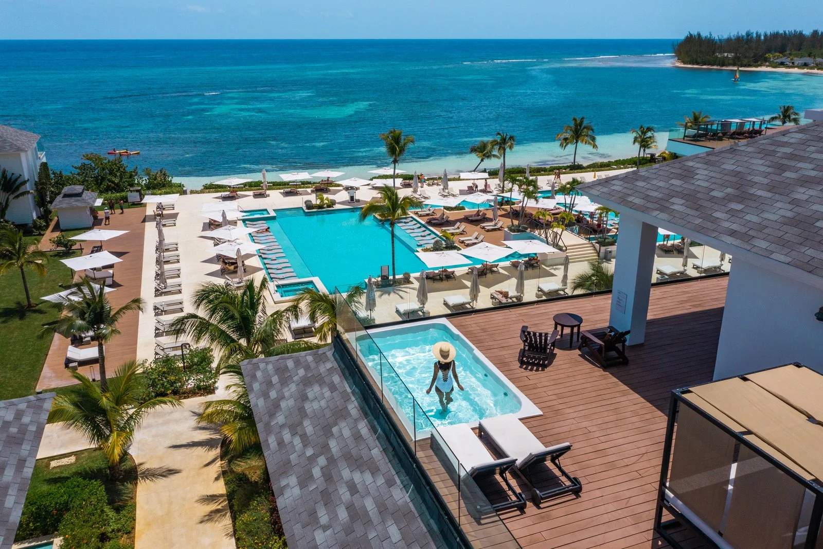 Woman in a hot tub on a balcony overlooking a pool and ocean at a tropical resort.