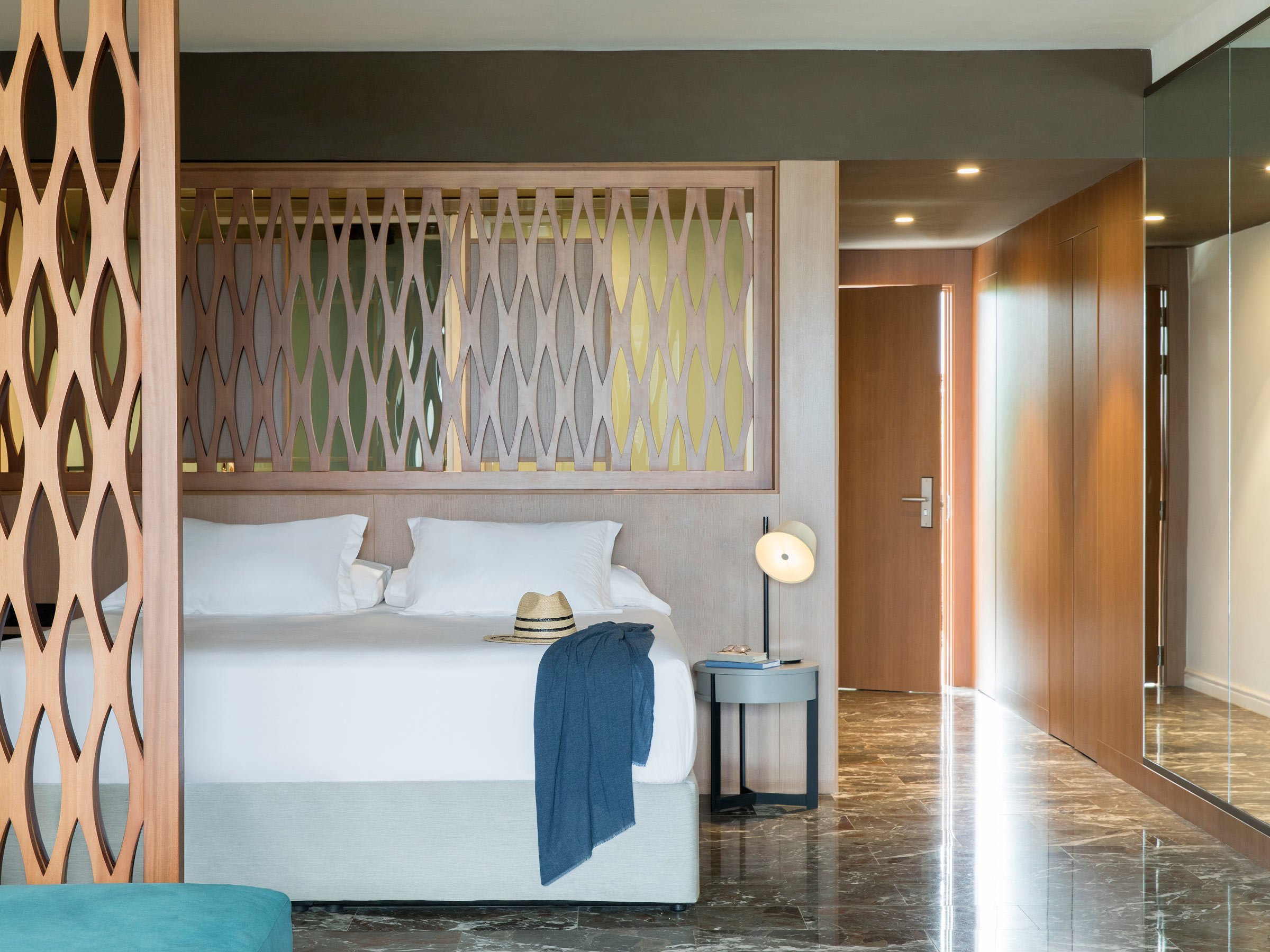 Modern hotel room with a bed protected by a wooden lattice partition, a straw hat and blue cloth on the bed, a small bedside table with a lamp, and a wooden door with light streaming in.