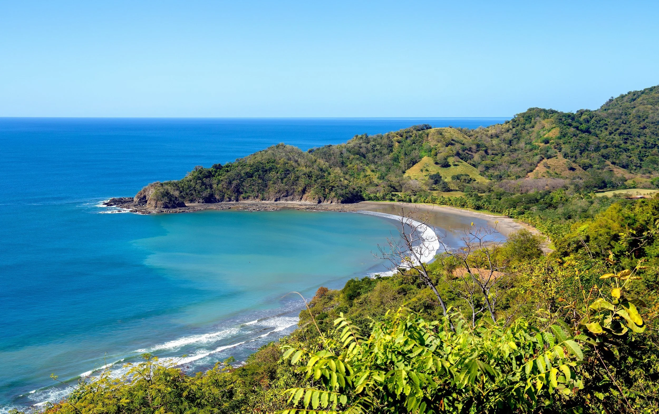 A scenic view of a tropical bay with blue water, a curved sandy beach, and green hills in the background under a clear sky.