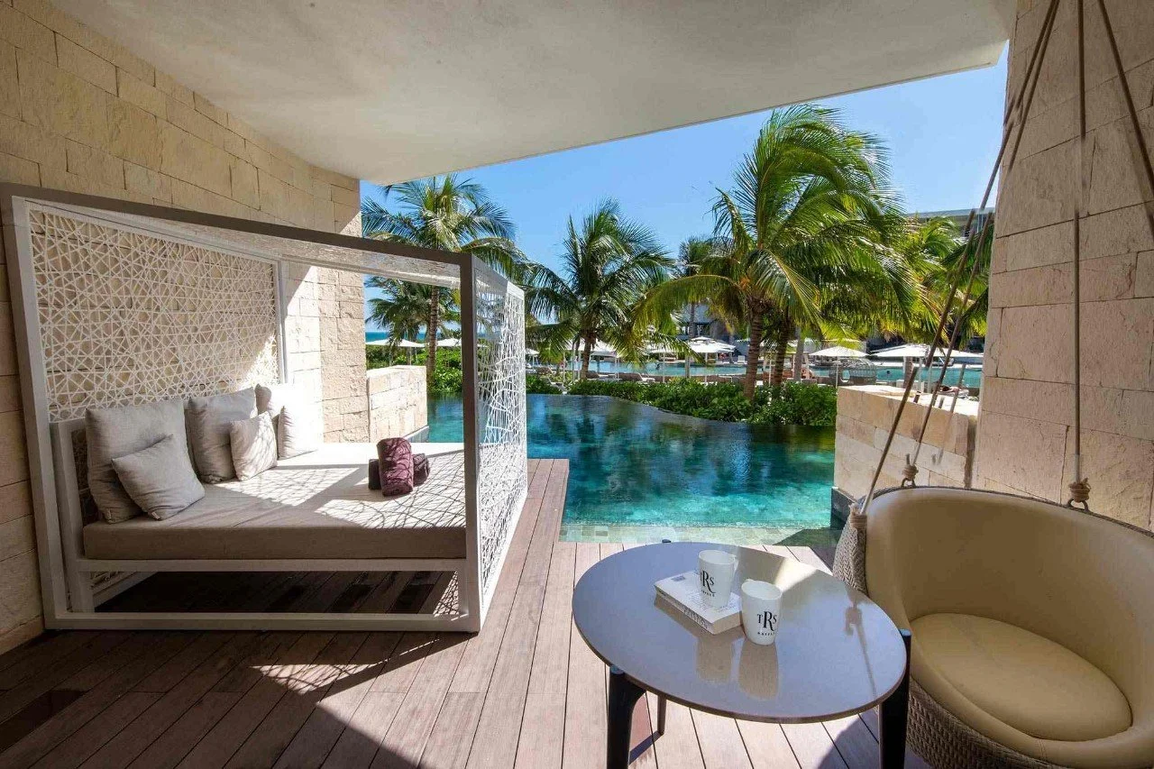 A luxurious poolside area with an outdoor daybed, a small table with cups, and lush palm trees in the background under a clear blue sky.