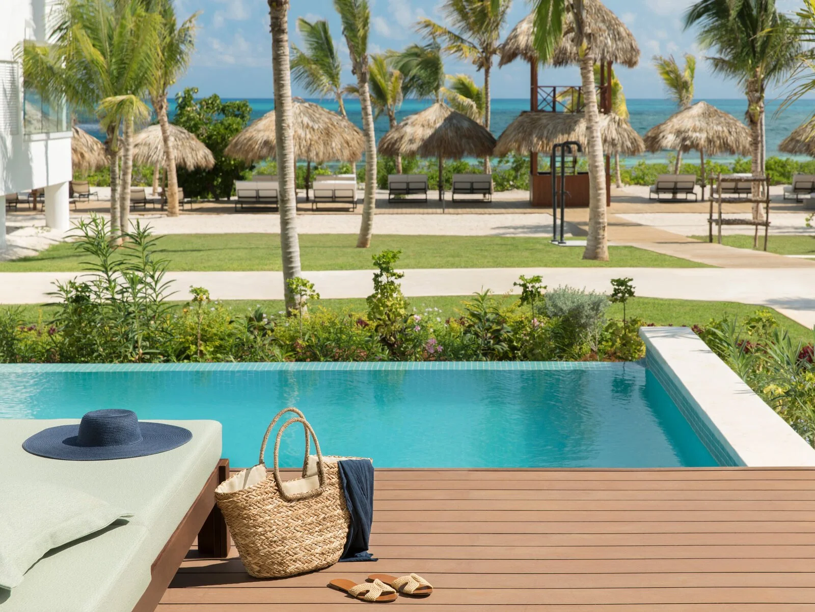 Poolside lounge chair with straw bag, hat, towel, and slippers next to a swimming pool, overlooking a tropical beach with palm trees and thatched roof cabanas.