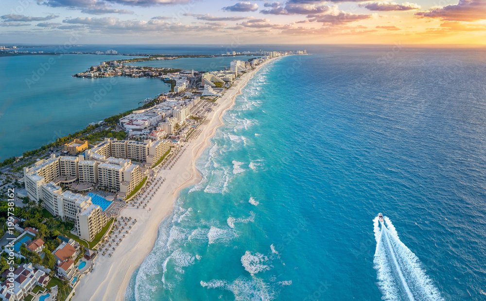 Aerial view of a beachside city with high-rise hotels along a sandy shoreline, with a boat cruising in the ocean, and a distant skyline at sunset.
