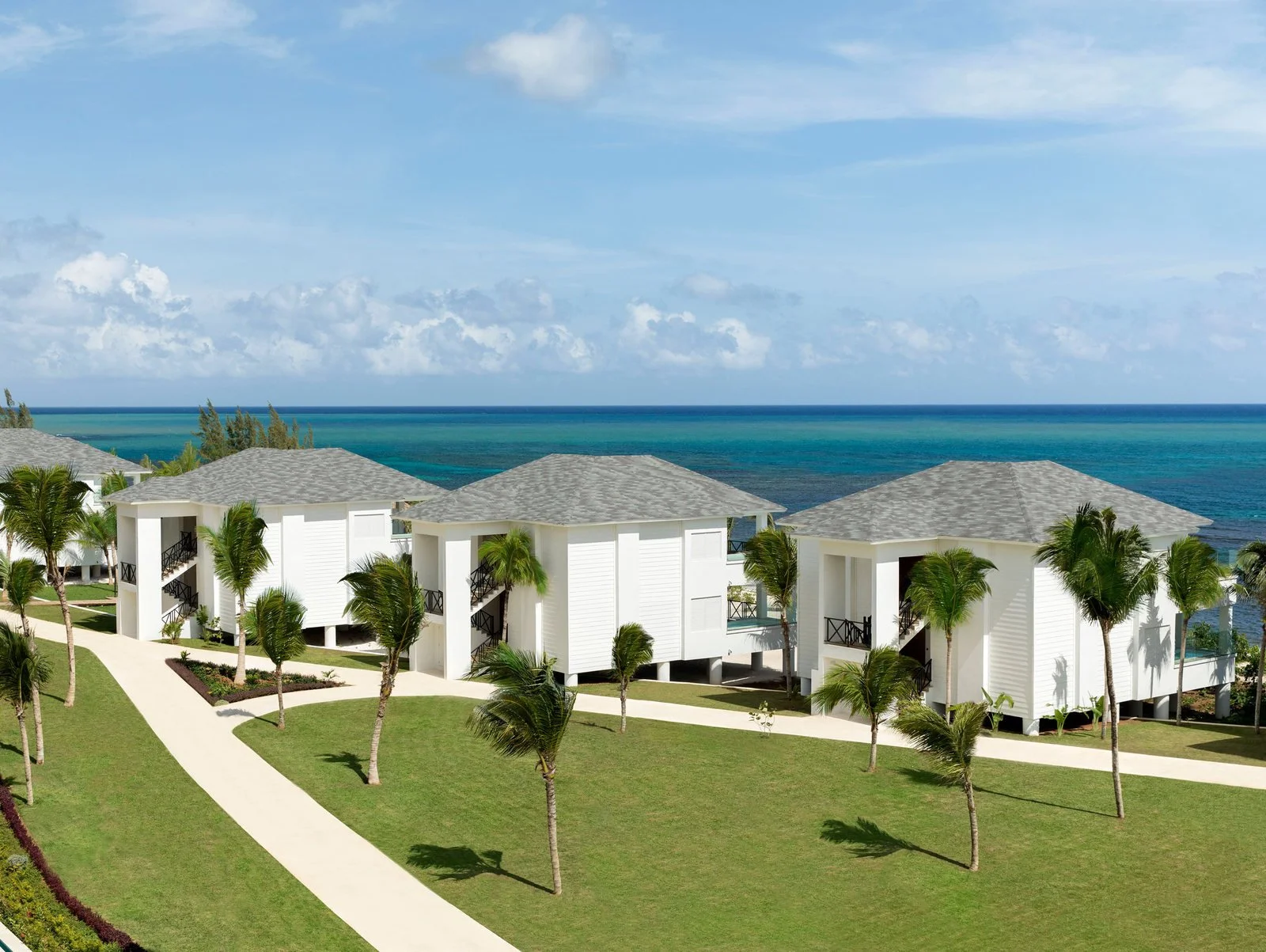 Beachfront resort with white buildings, grey roofs, and palm trees on grassy lawns, overlooking the ocean under a partly cloudy sky.