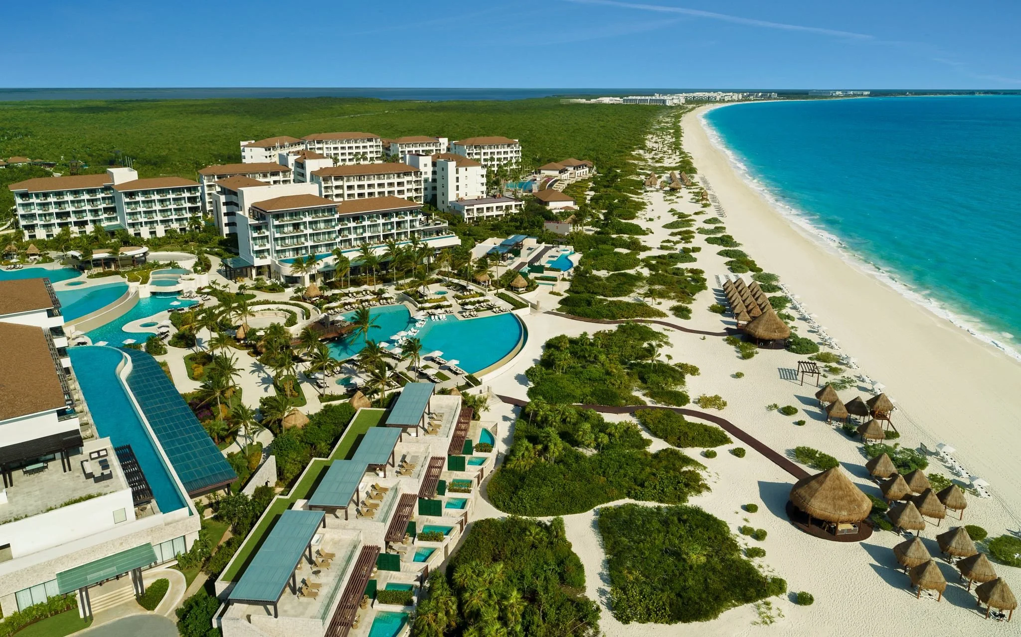 Aerial view of a luxurious beach resort with multiple swimming pools, white sandy beach, thatched beach huts, and green vegetation along the shoreline.