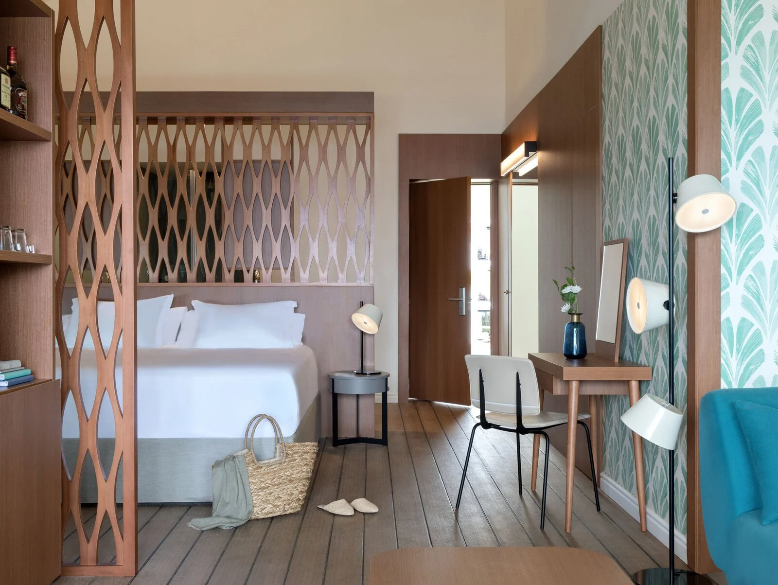 Hotel room with a bed, nightstand, and desk with a chair. Decorated with a blue and green leaf patterned wallpaper and wooden accents. Includes a woven basket with a towel, slippers, and a tote bag.