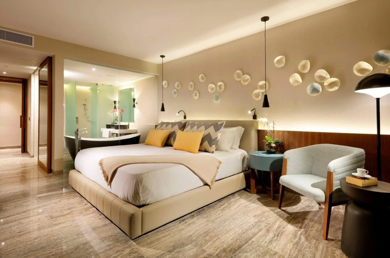 A modern hotel room with a large bed, decorative pillows, a side table with a plant, a reading chair, a floor lamp, and a wall installation of round decorative objects. There is a visible bathroom area with a bathtub in the background.