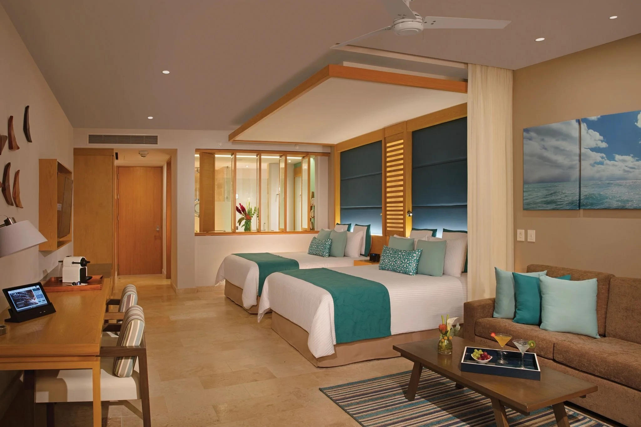 A modern hotel room with two beds, a sofa with teal pillows, a wooden table with drinks and snacks, a desk with a tablet, and a wall-mounted TV. There are decorative elements like artwork and a large window with a view of the ocean.